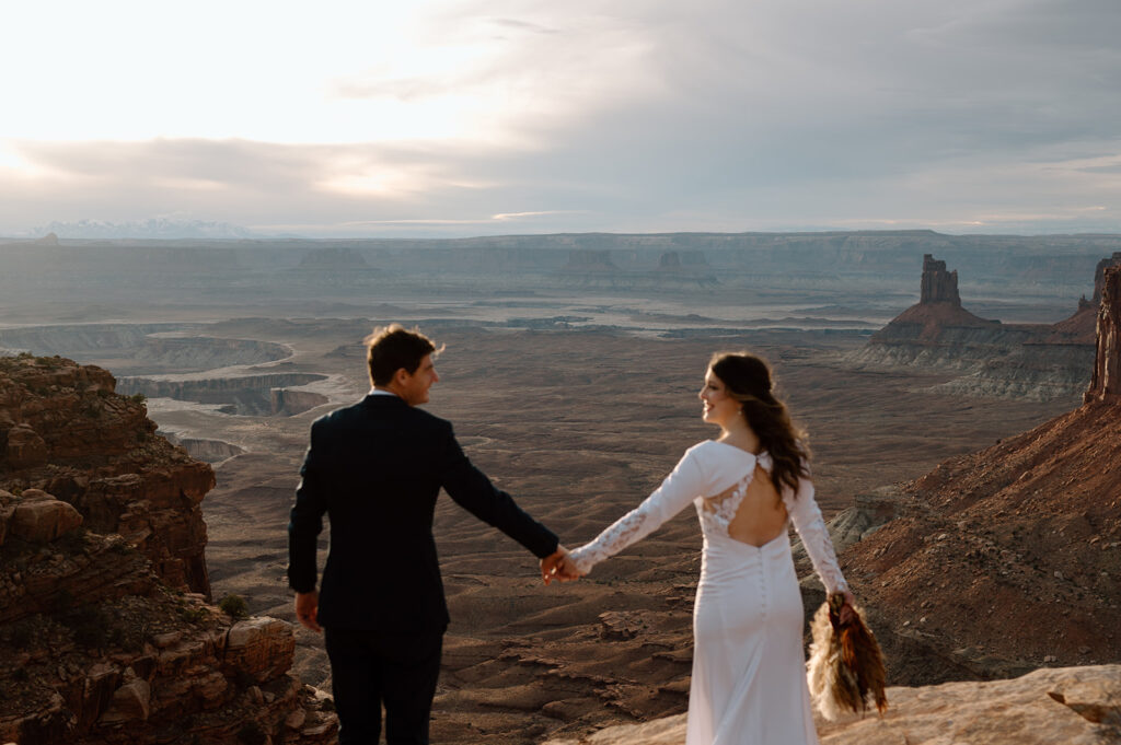 A Utah elopement couple dressed in formal wedding attire looks at one another while holding hands on an overlook in Canyonlands National Park. 