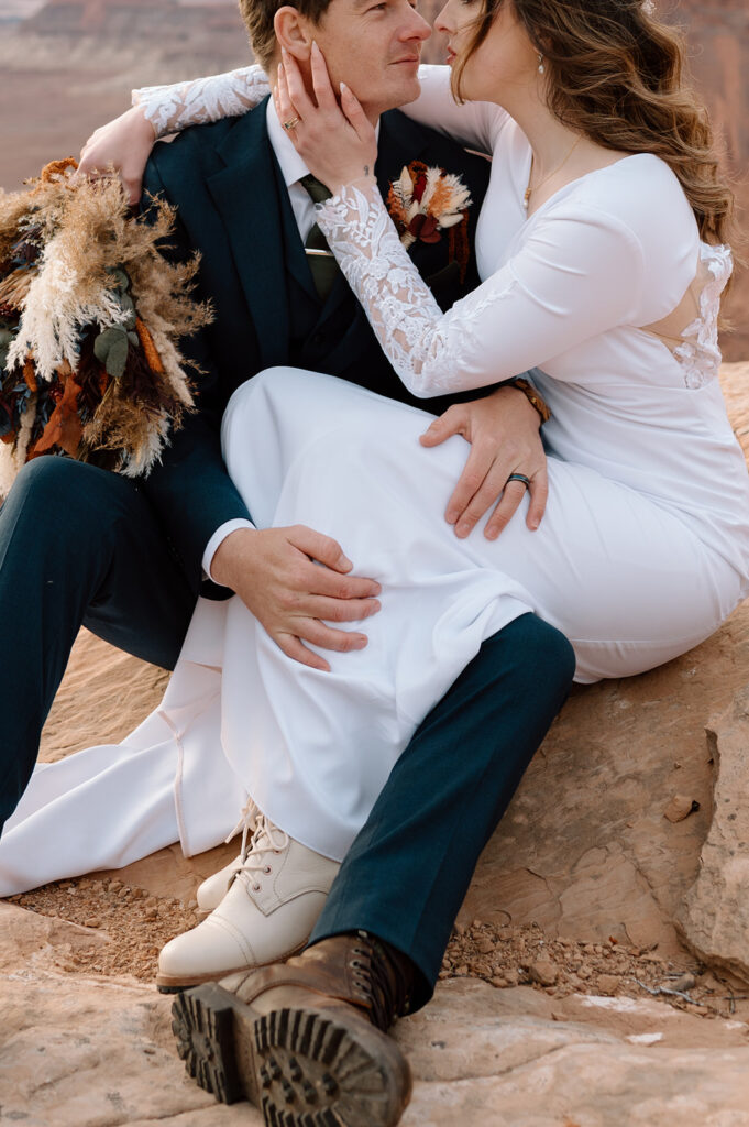 A groom in a navy tuxedo, green tie, and brown boots, sits on a large red rock with his bride, who is wearing a long white wedding dress and cream boots. 