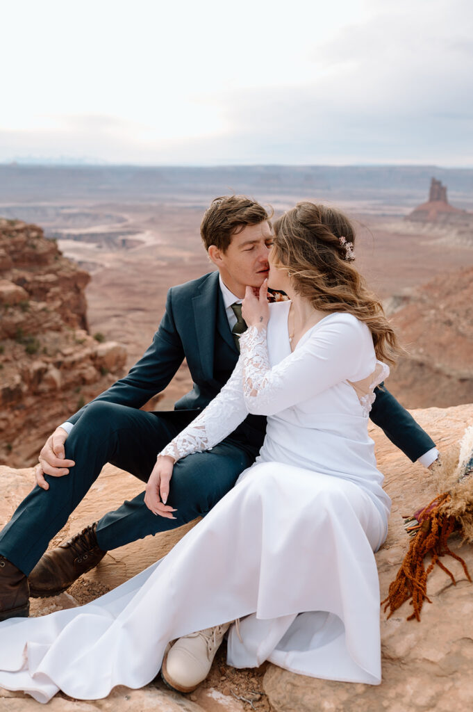 A bride kisses her groom while sitting on a red rock that overlooks Canyonlands National Park.