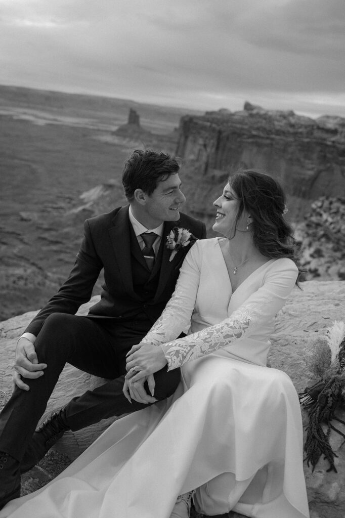 A bride and groom sit with one another on a red rock at Grand View Point Overlook in Canyonlands. 