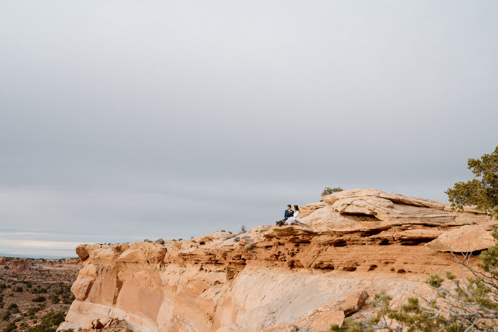 A Utah elopement couple sits on a cliff's edge and watches the sun set over Canyonlands National Park. 