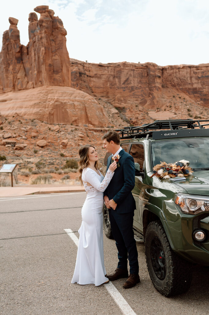 A woman wearing a long white wedding dress adjusts her groom's dried floral boutonniere near a 4x4 vehicle in Arches National Park. 