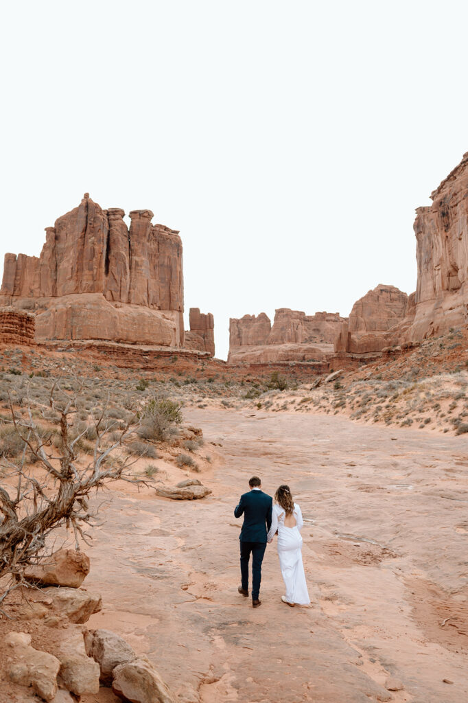 A desert elopement couple in formal wedding attire walks along red rock slabs in Moab. 