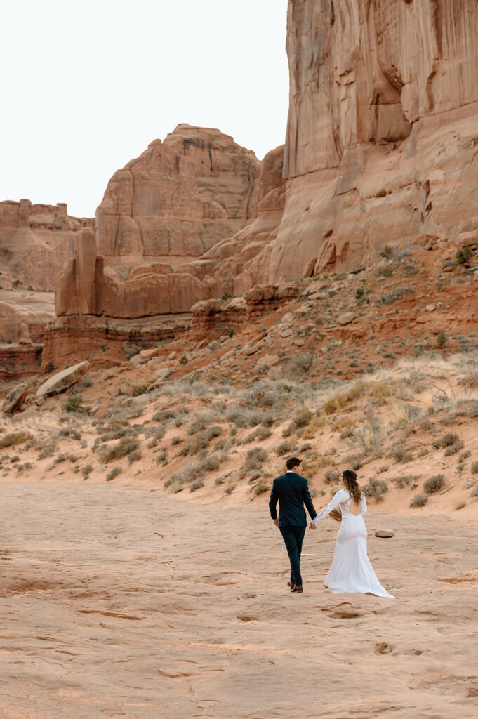 An adventure elopement couple in formal wedding attire walks along red rocks in Arches National Park. 