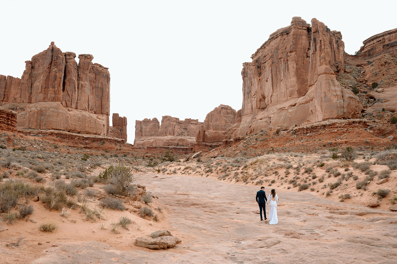 A Utah elopement couple holds hands while walking towards large rock formations in the Moab desert.