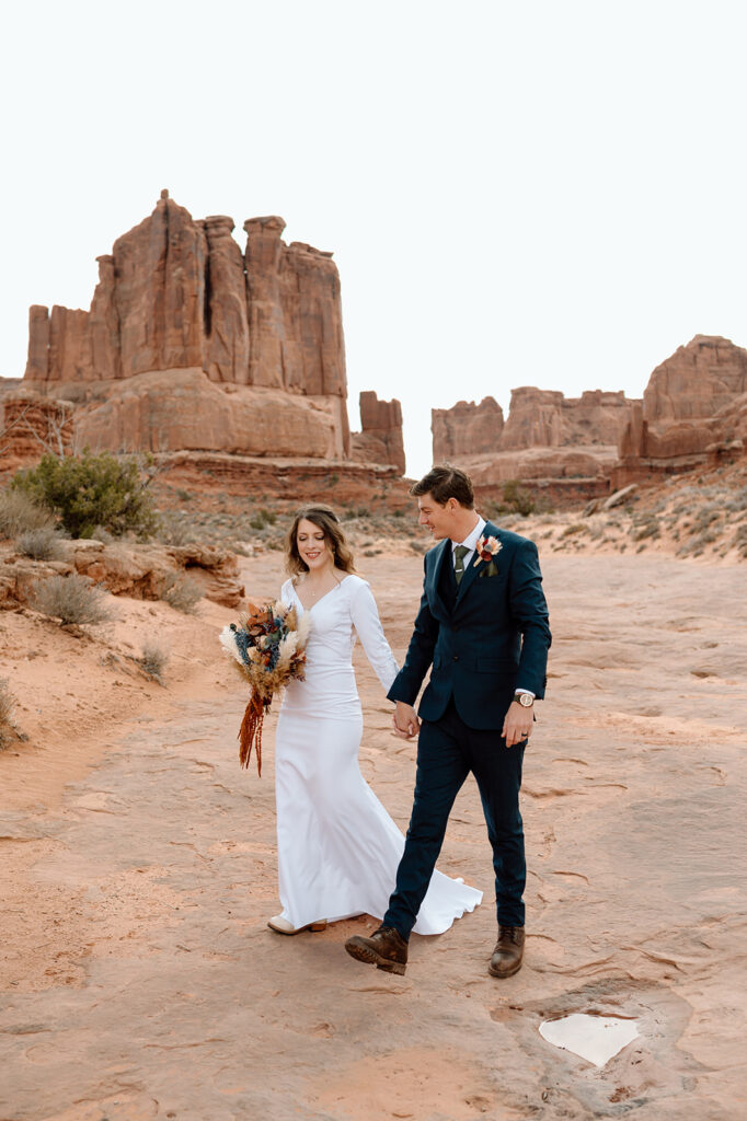 A couple holds hands and walks along a path in Arches National Park during their adventure elopement in Moab, Utah. 