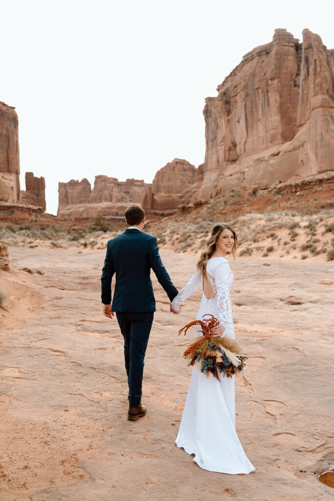 A bride looks over her shoulder while holding a dried bridal bouquet and her grooms hand in Arches National Park. 