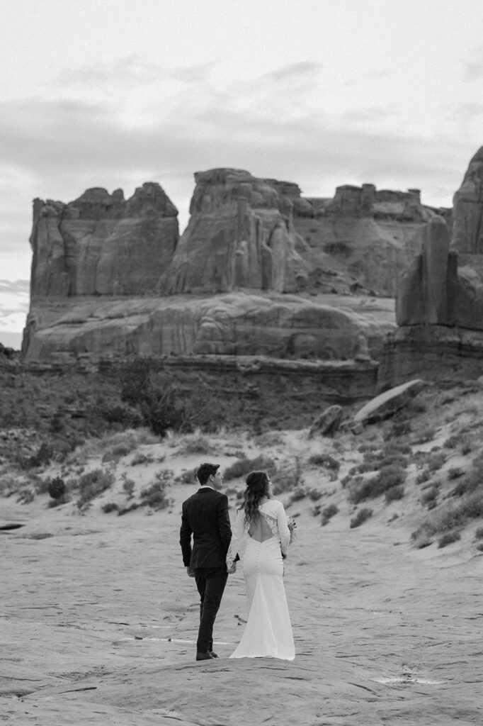 A couple walks along red rocks while holdings hands during their Arches National Park elopement. 