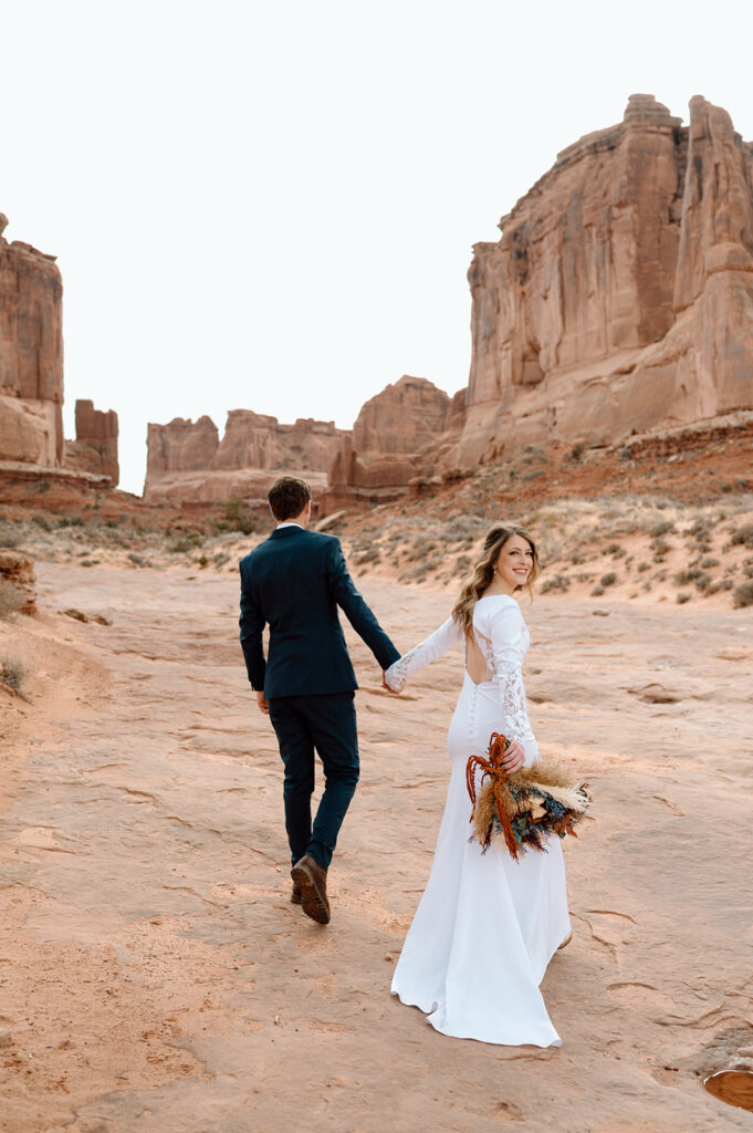 A couple holds hands as a bride looks over her shoulder wearing a white wedding dress and holding a dried floral bouquet. 