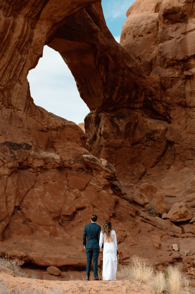 A couple admires a red rock arch in Arches National Park during their Moab elopement. 