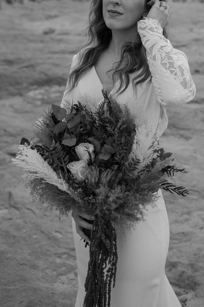 A bride holds a dried bridal bouquet, while wearing a long wedding dress with lace sleeves. 