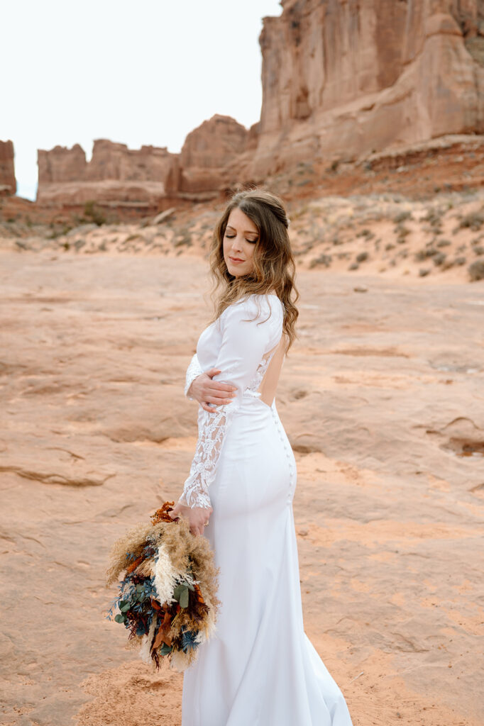 A woman in a long sleeve wedding dress holds a dried floral bouquet in Arches National Park. 