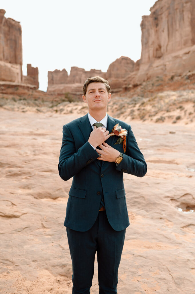 A groom, wearing a navy tuxedo and dried floral boutonniere, adjusts his tie while standing on red rocks in Arches National Park. 