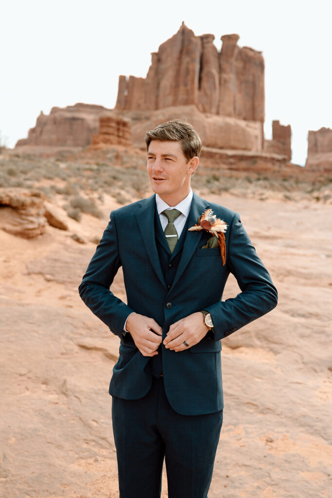 A man stands near rock structures in Arches National Park in a navy suit and buttons his jacket that is adorned with a dried floral boutonniere on the jacket's left lapel. 